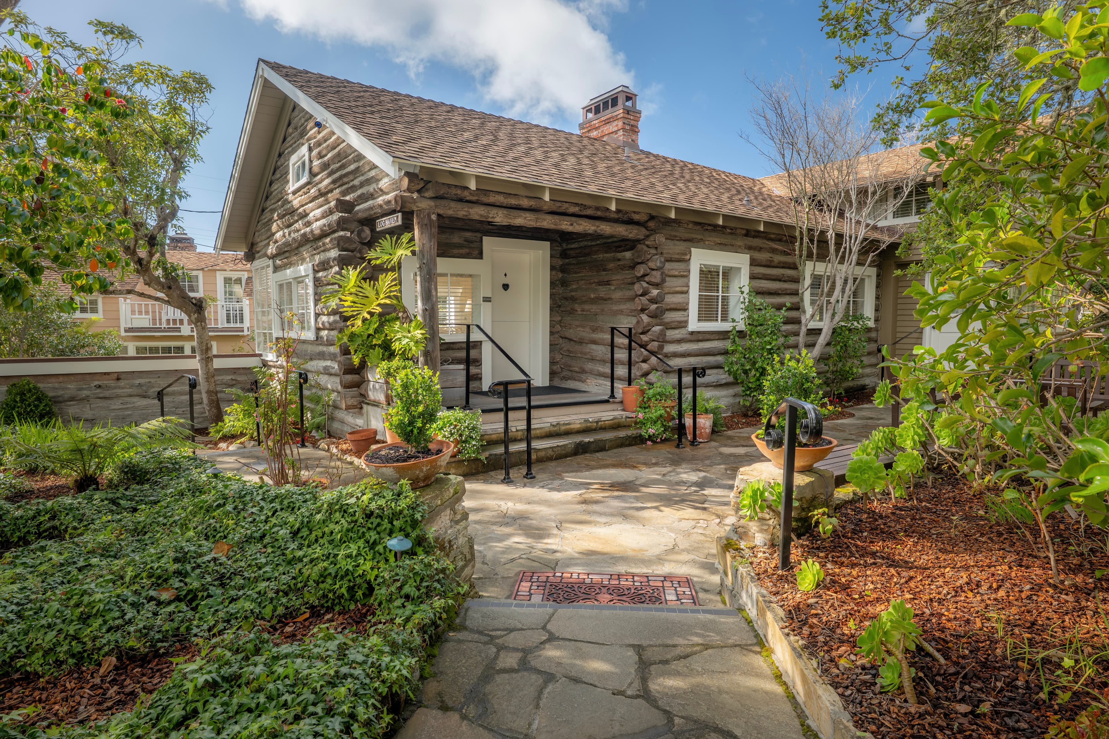 Exterior view of Log Haven, a historic log cabin with a brick chimney and large stone patio entrance, surrounded by lush green garden beds and trees under a clear blue sky.