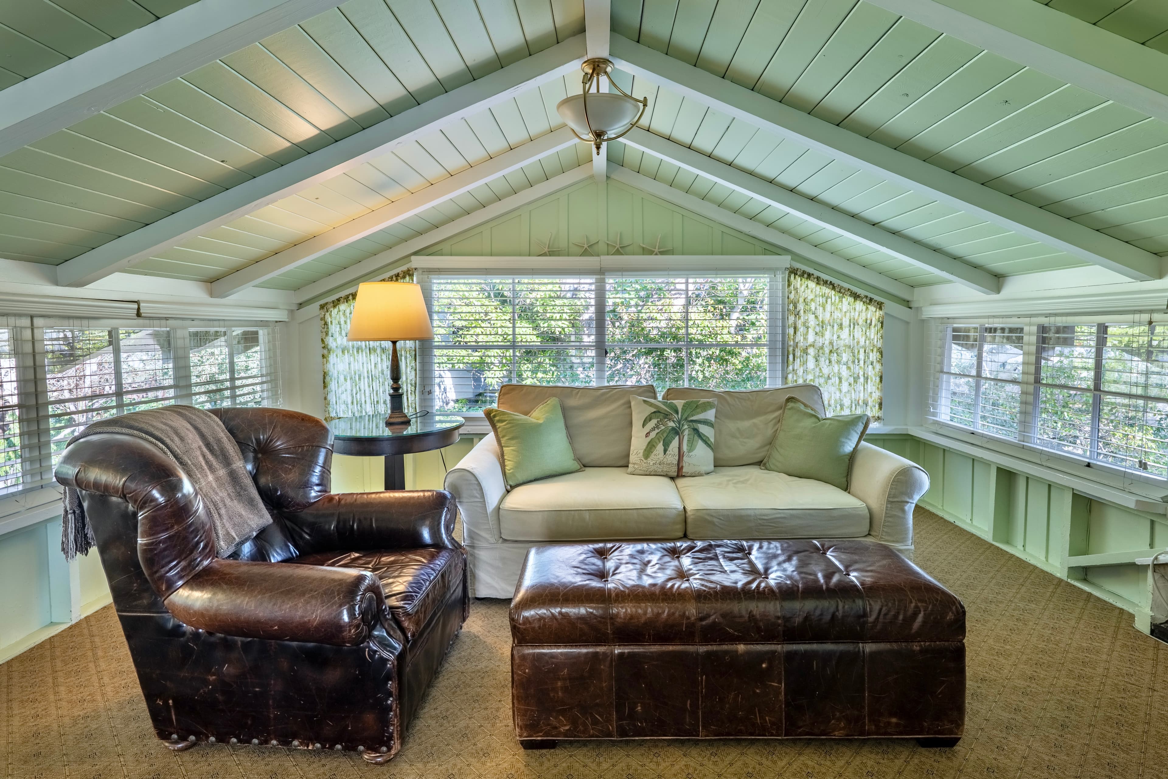 A cozy upstairs living area with a vaulted white-beamed ceiling, featuring a large leather armchair and tufted ottoman in front of a neutral-toned sofa with green accent pillows.