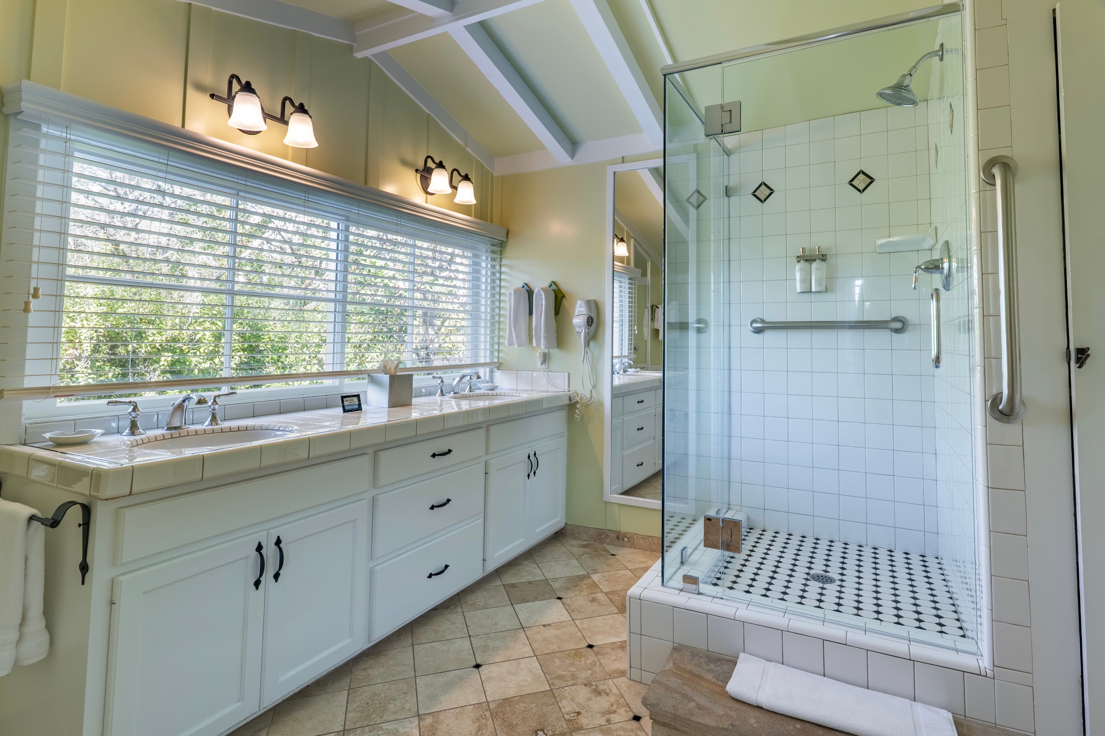 A bright, spacious bathroom with light green walls and a white-beamed ceiling, featuring a wide vanity with double sinks next to a large glass-enclosed walk-in shower with a safety grab bar.