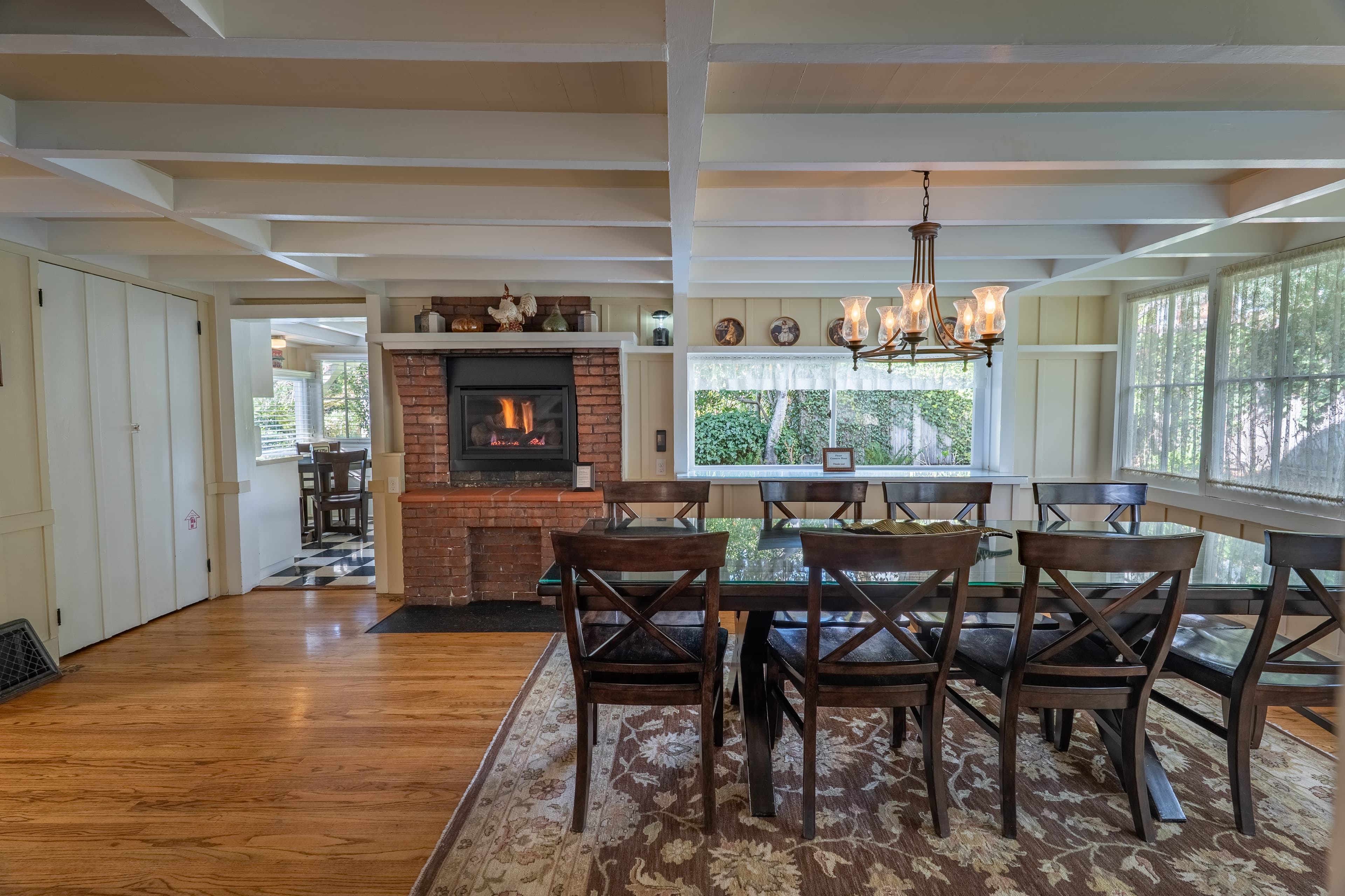 A spacious dining room with a long dark wood table and cross-back chairs, featuring a brick fireplace with black insert, a glowing chandelier, and white- beamed ceilings.