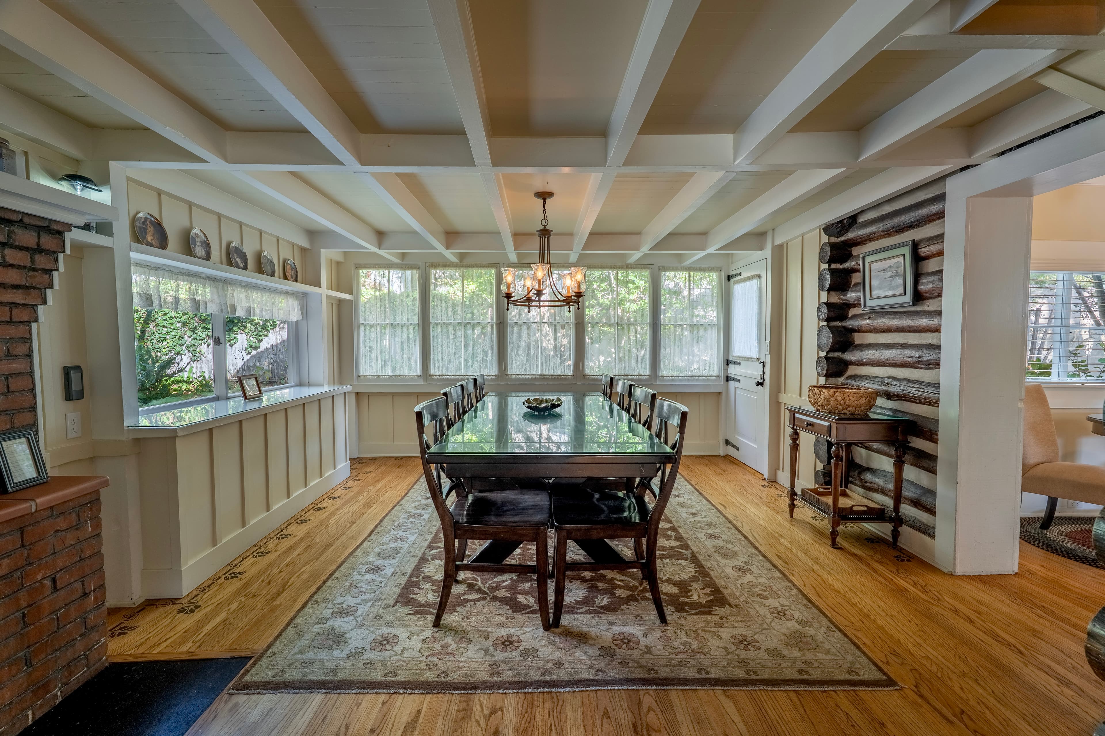 A long glass-topped dining table, featuring stenciled wood floors, white-beamed ceilings, and a view of a rustic log wall.