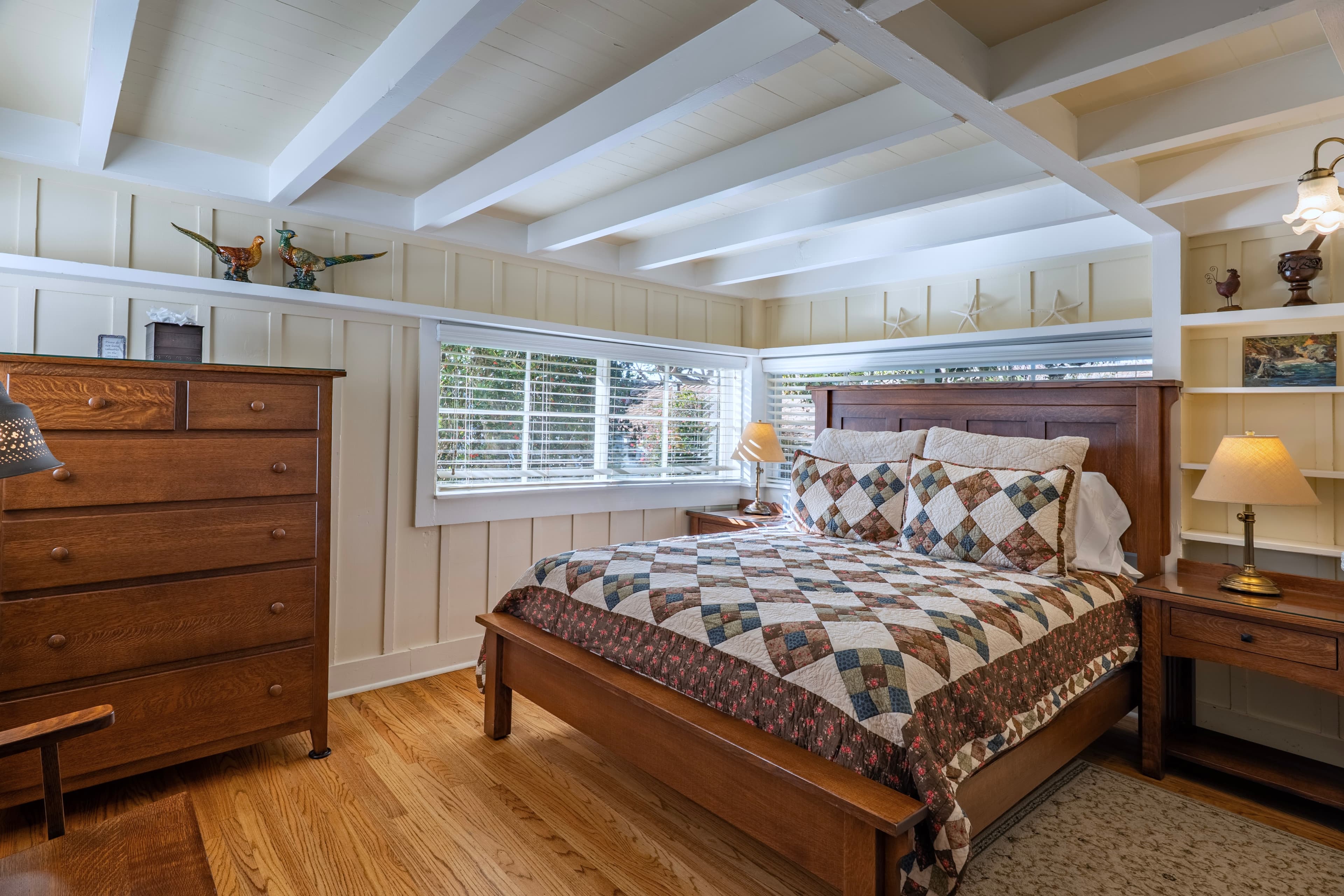 A cozy bedroom in the Log Haven cabin featuring wood floors, a large dark wood bed with a patterned quilt, and a matching tall dresser against wood-paneled walls under a white-beamed ceiling.