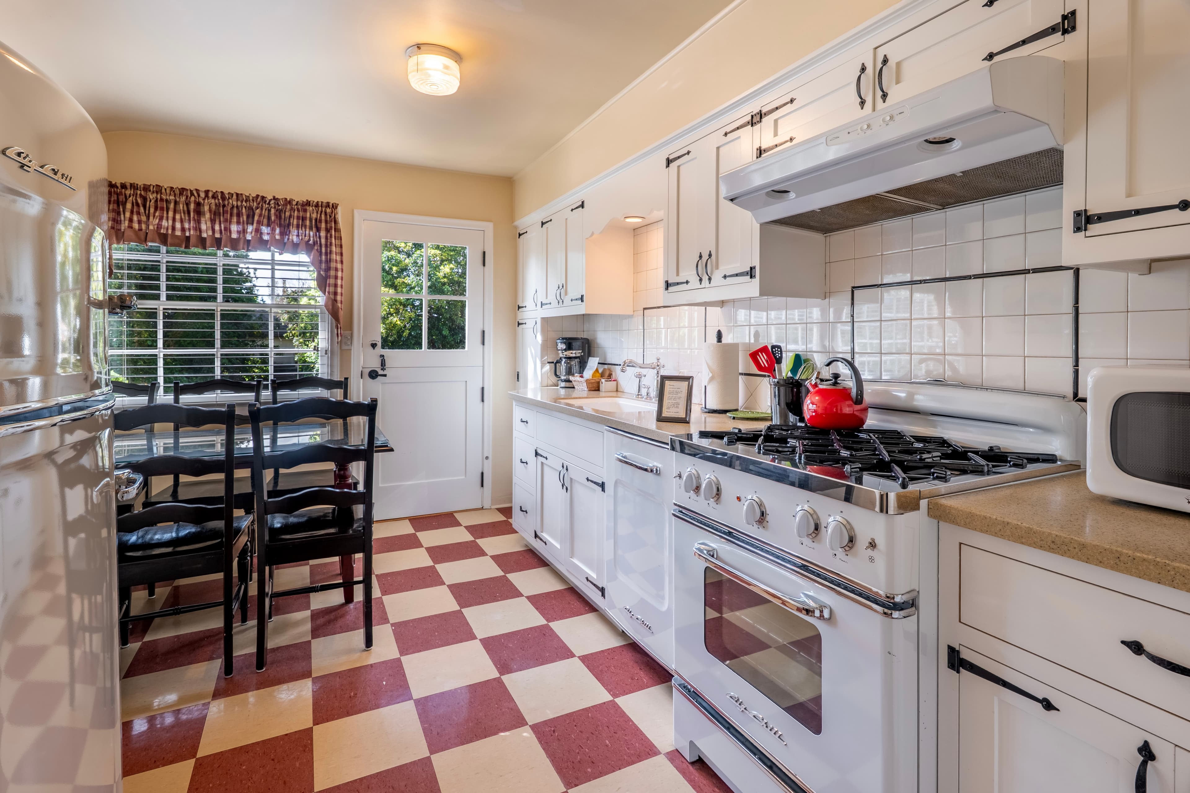 A vintage-style kitchen with a red and white checkered floor, featuring a large white retro gas range, white cabinets with black iron latches, and a dark dining table with six chairs next to a window.