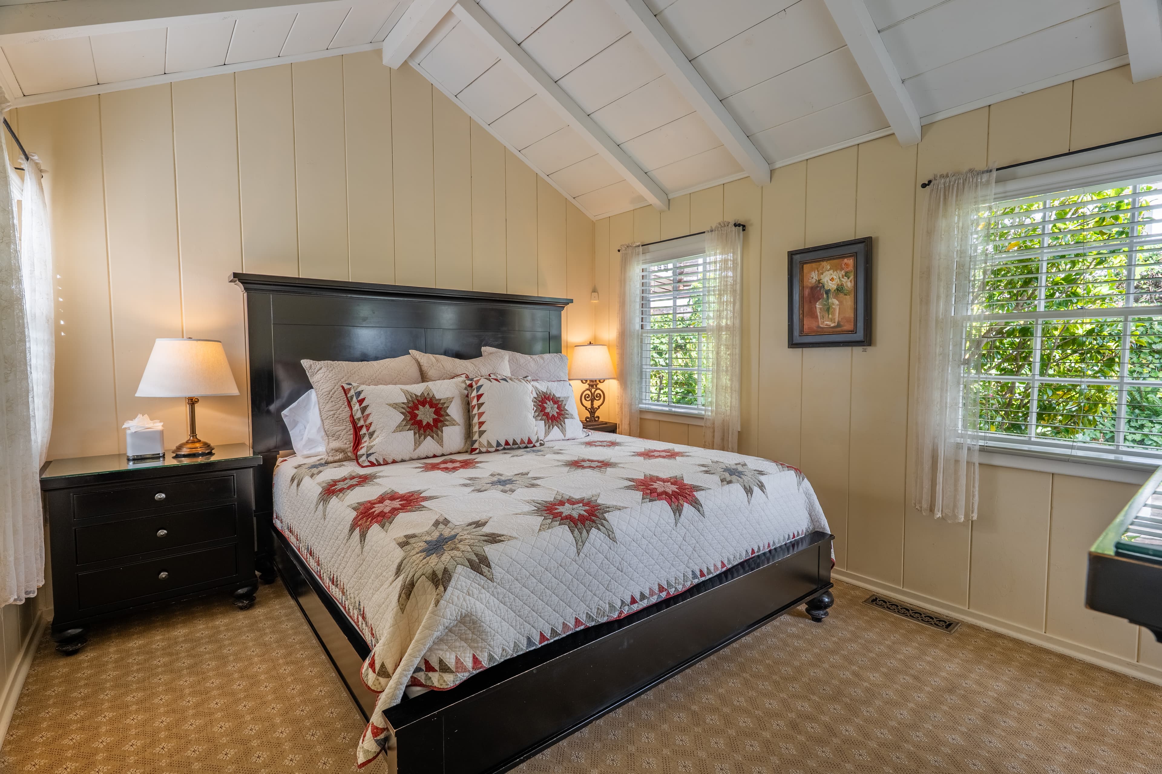 A bright bedroom with cream-colored wood-paneled walls and a white vaulted white-beamed ceiling, featuring a large dark wood bed with a patterned quilt and bedside table.