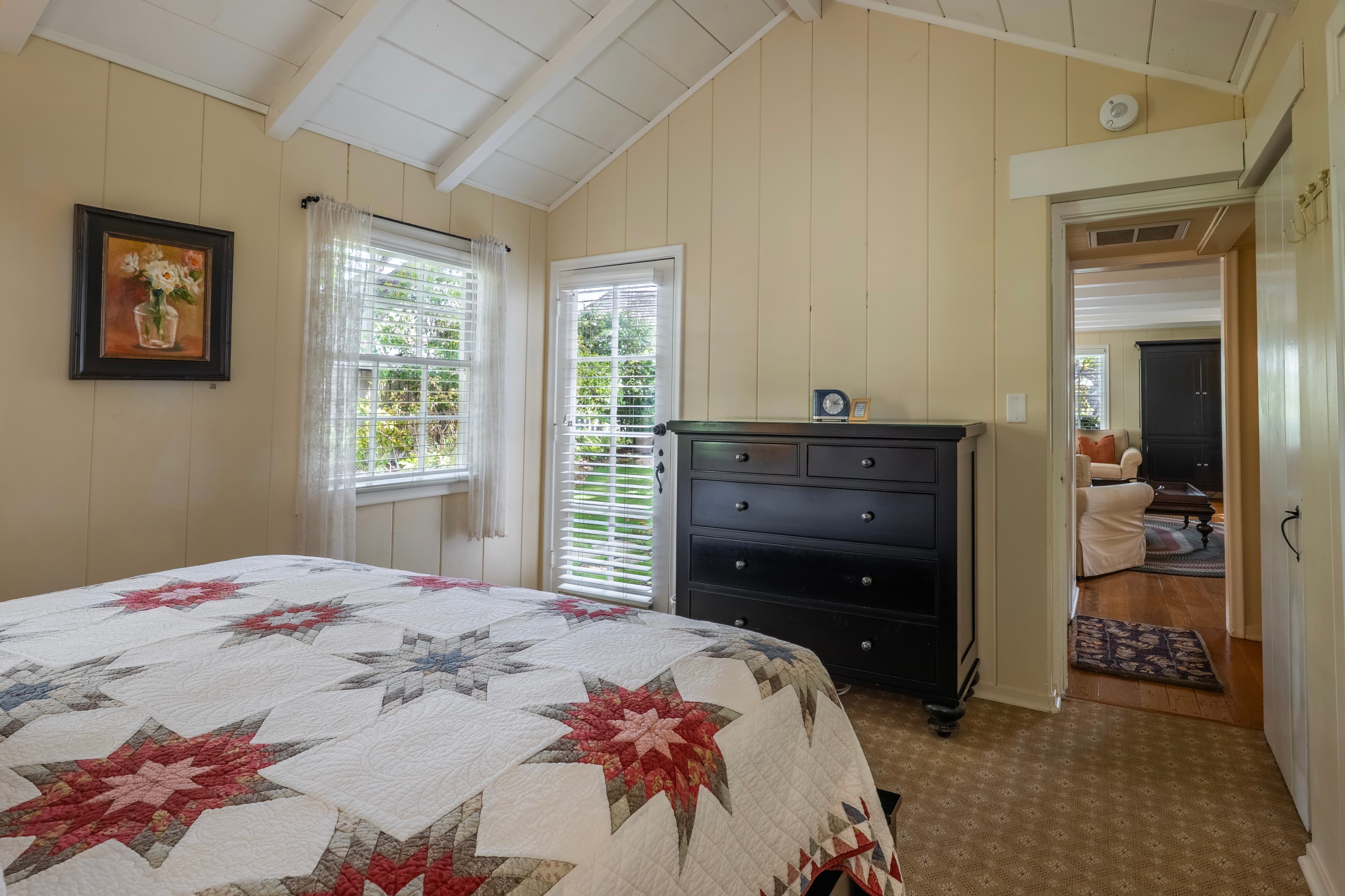 A bright, cozy bedroom with cream-colored wood-paneled walls and a vaulted white-beamed ceiling, featuring a large dark wood bed covered in a patterned quilt.