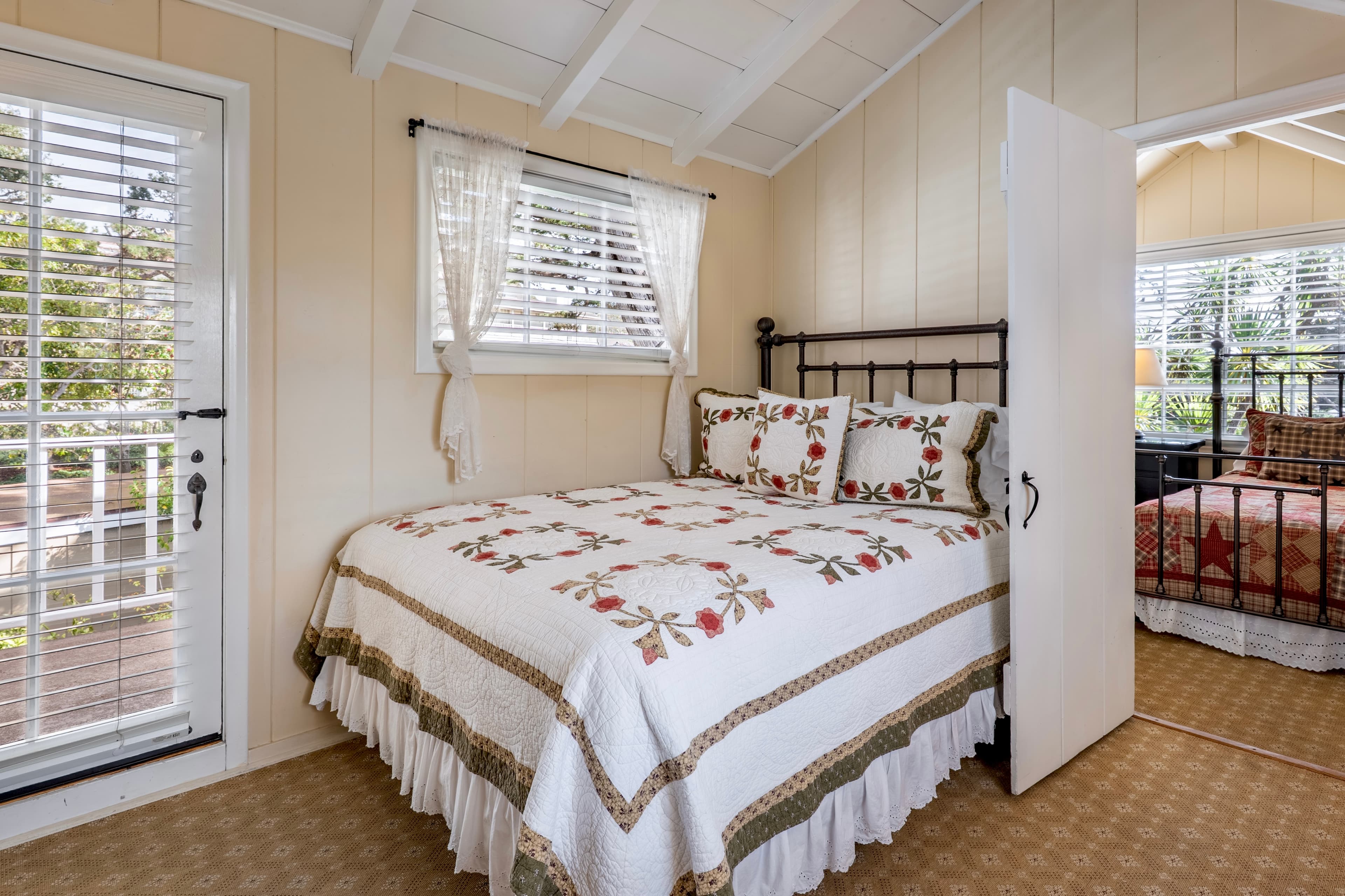 A cozy cottage bedroom with light cream walls and white-beamed vaulted ceilings, featuring a bed with a patterned quilt and a glass door leading to a balcony.