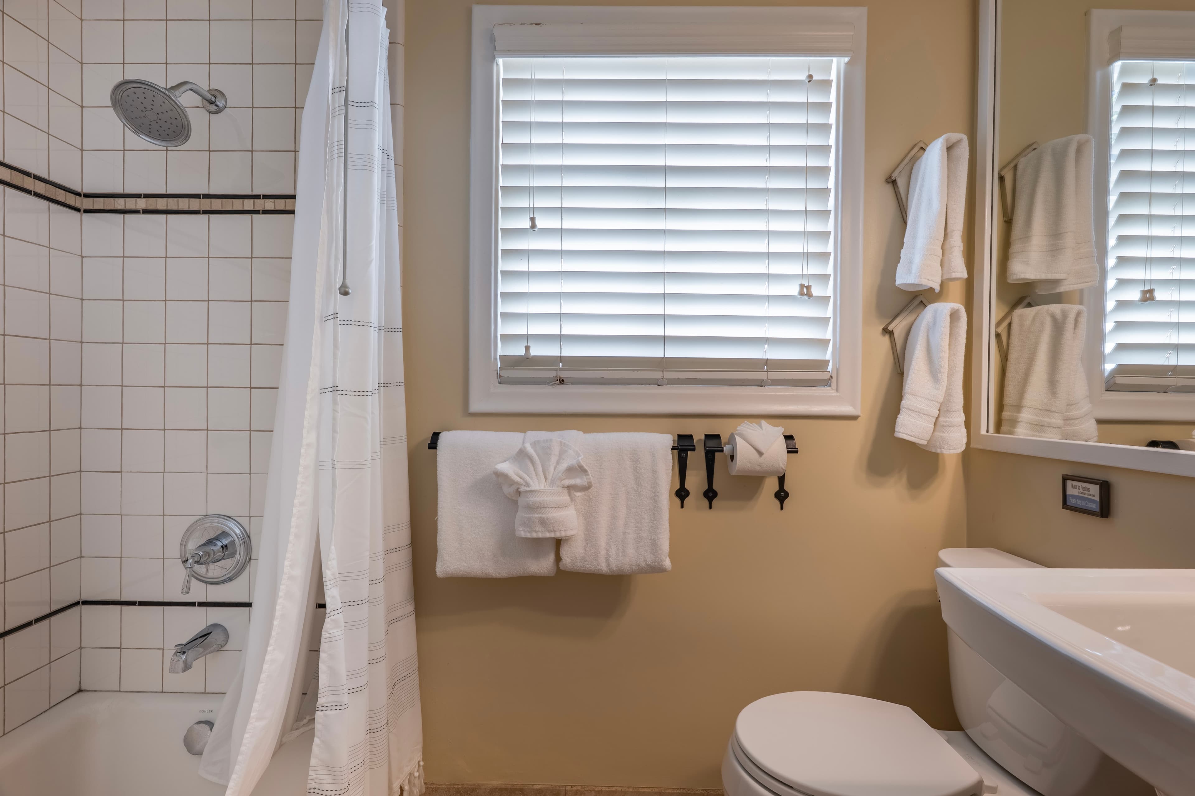 A classic bathroom with cream walls and white tile, featuring a white pedestal sink, a toilet, and a bathtub with a white shower curtain and a chrome showerhead.