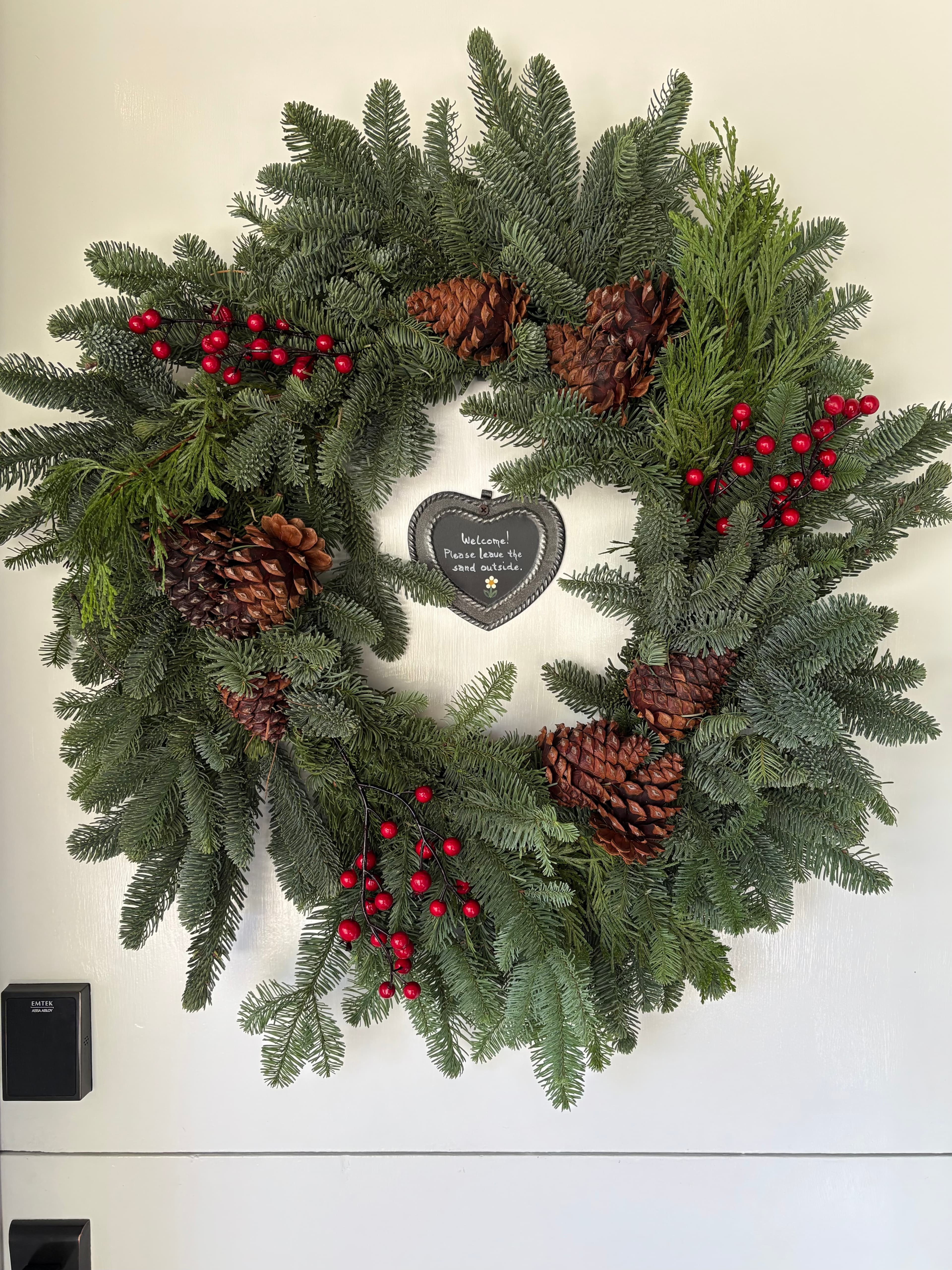 For December guests, an evergreen wreath decorated with pinecones and red berries, hanging on a white door with a small heart-shaped sign in the center.