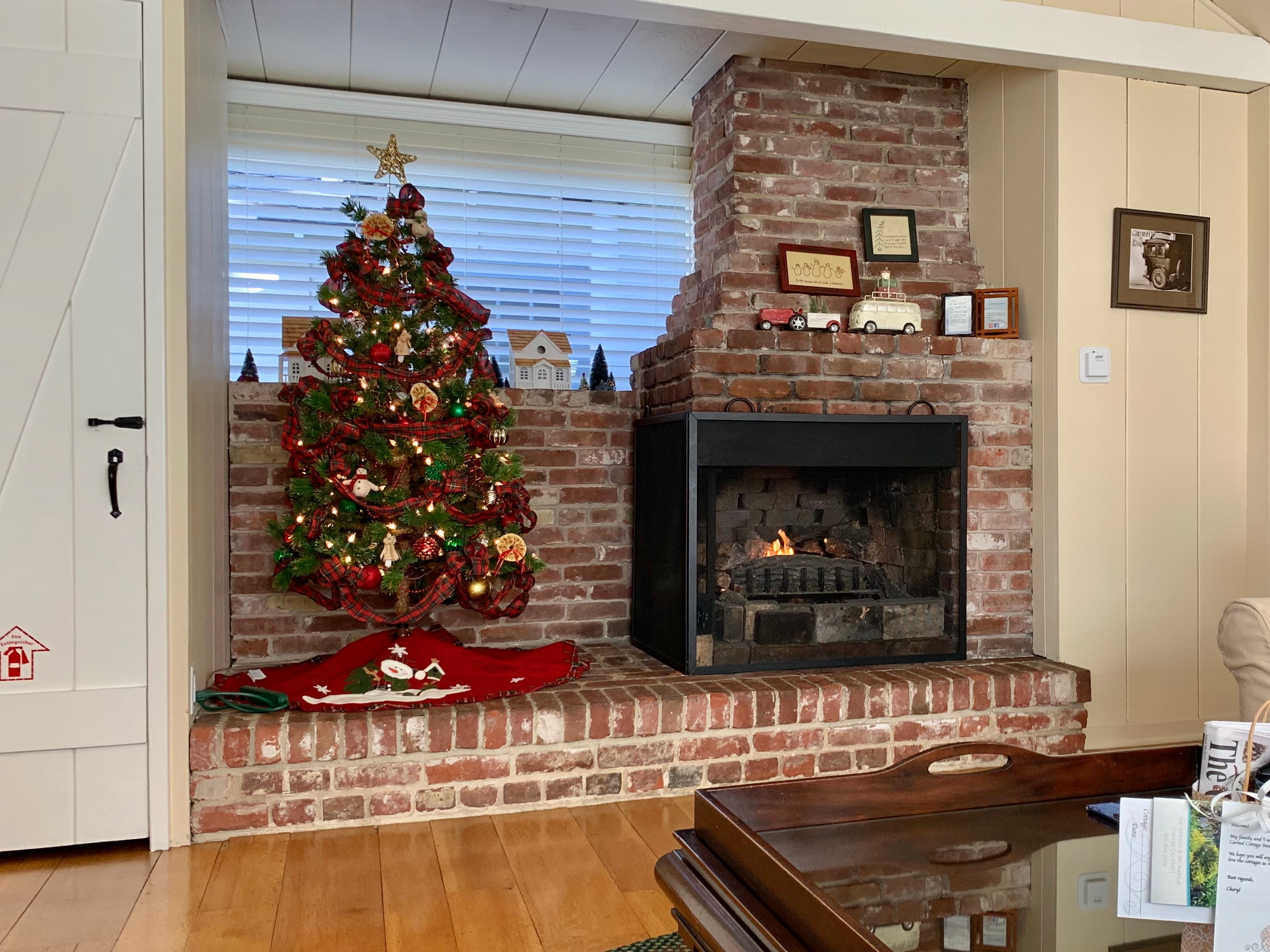 For December guests, a cozy living room during the holidays, featuring a decorated Christmas tree with a red tree skirt and a lit fireplace set within a large brick hearth.