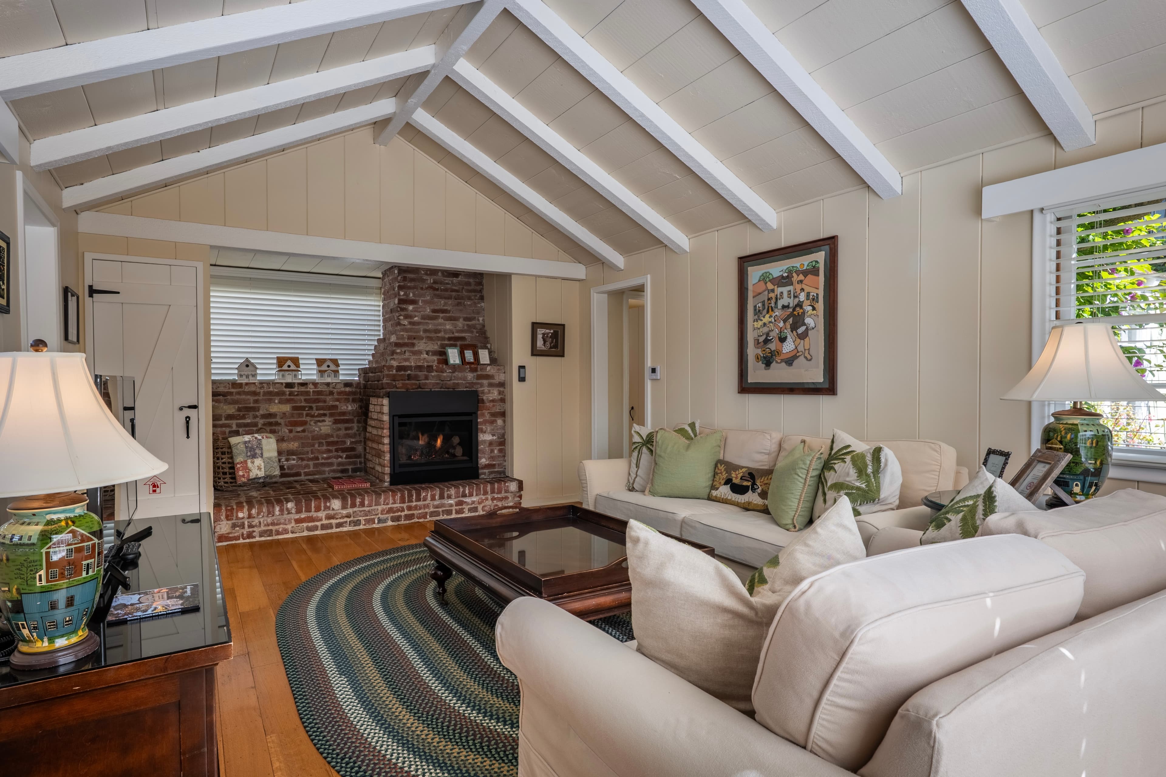 A cozy living room with a vaulted white-beamed ceiling and hardwood floors, featuring a brick, black insert fireplace, neutral-toned sofas with green accent pillows, and a large oval area rug.