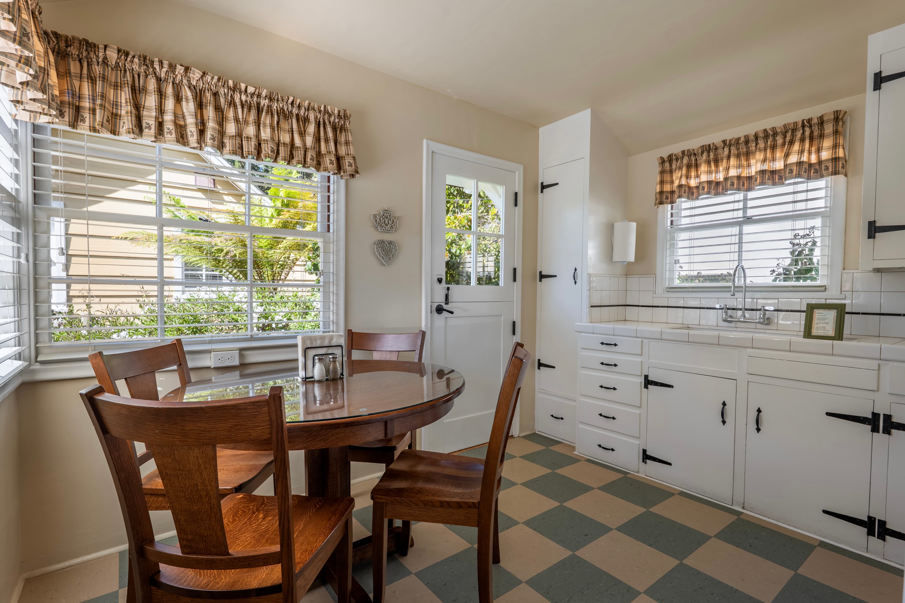 A bright dining nook and kitchen featuring a brown wooden circular table with four chairs, set on a tan and green checkered floor next to large windows with plaid valances.