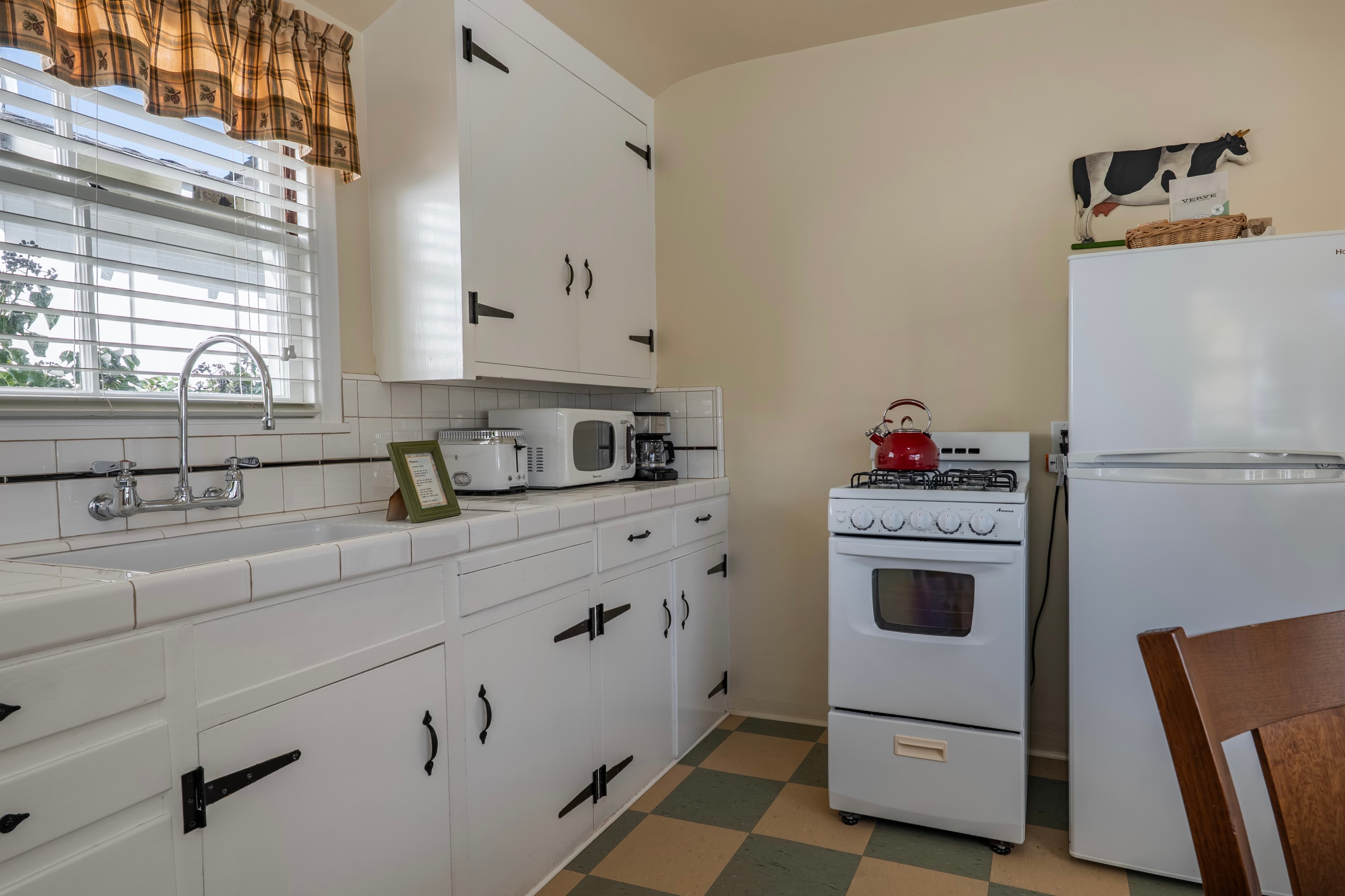 A classic kitchen with a tan and green checkered floor, featuring white shaker-style cabinets with black rustic latches, a white gas stove, and a small white refrigerator.
