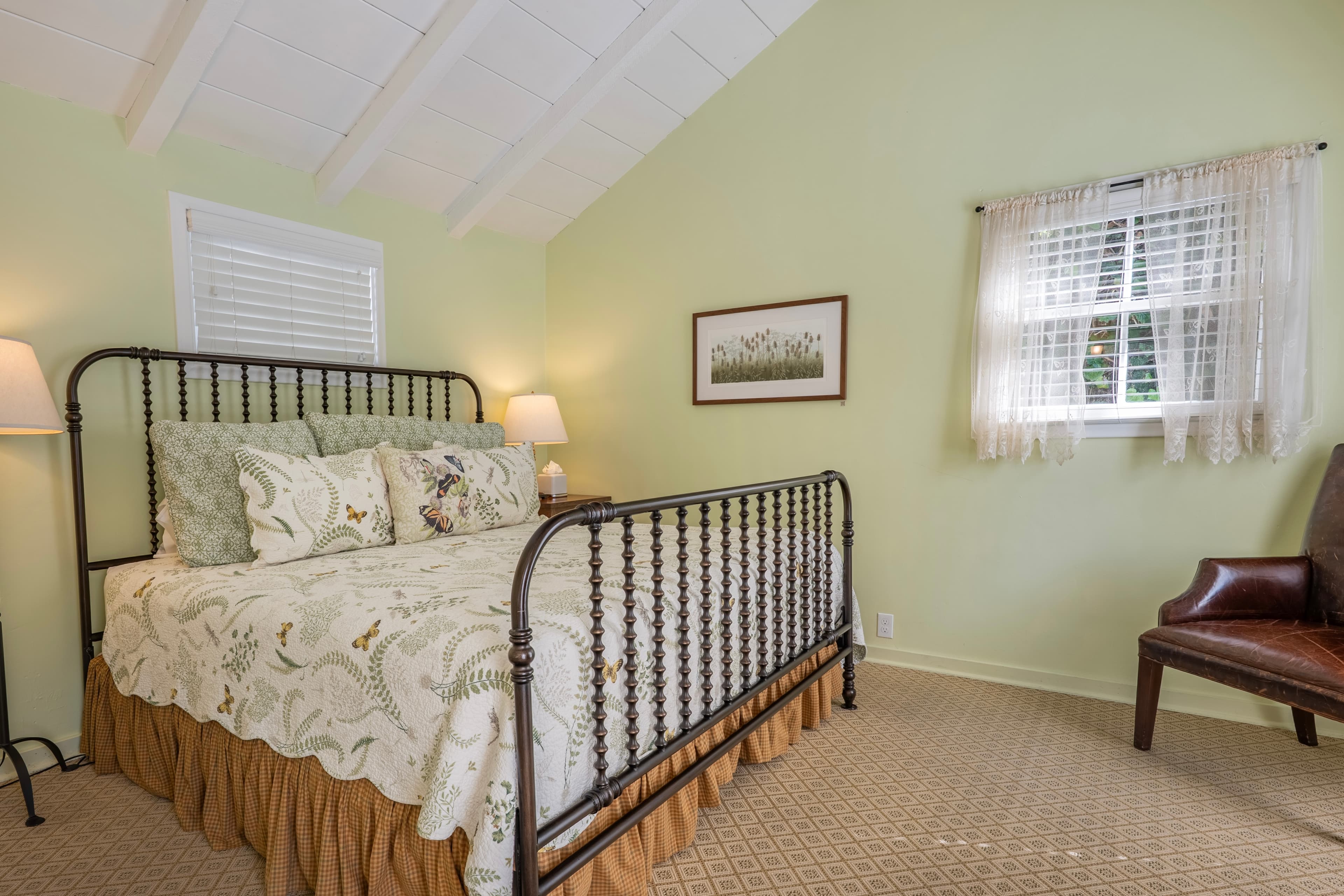 A bright bedroom with pale green walls and a vaulted white-beamed ceiling, featuring a dark metal bed frame with a patterned quilt and a brown leather armchair.
