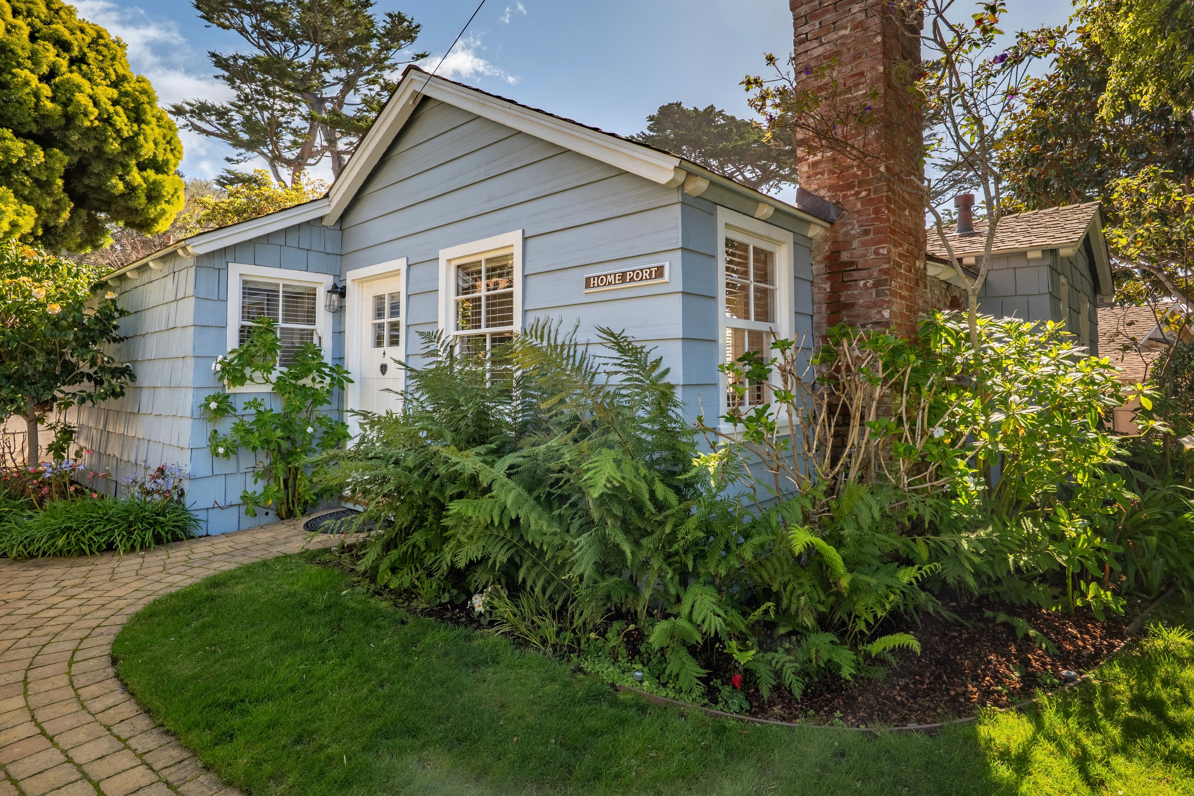 A blue cottage with a white door, surrounded by a lush garden and a winding paver path under a clear blue sky.