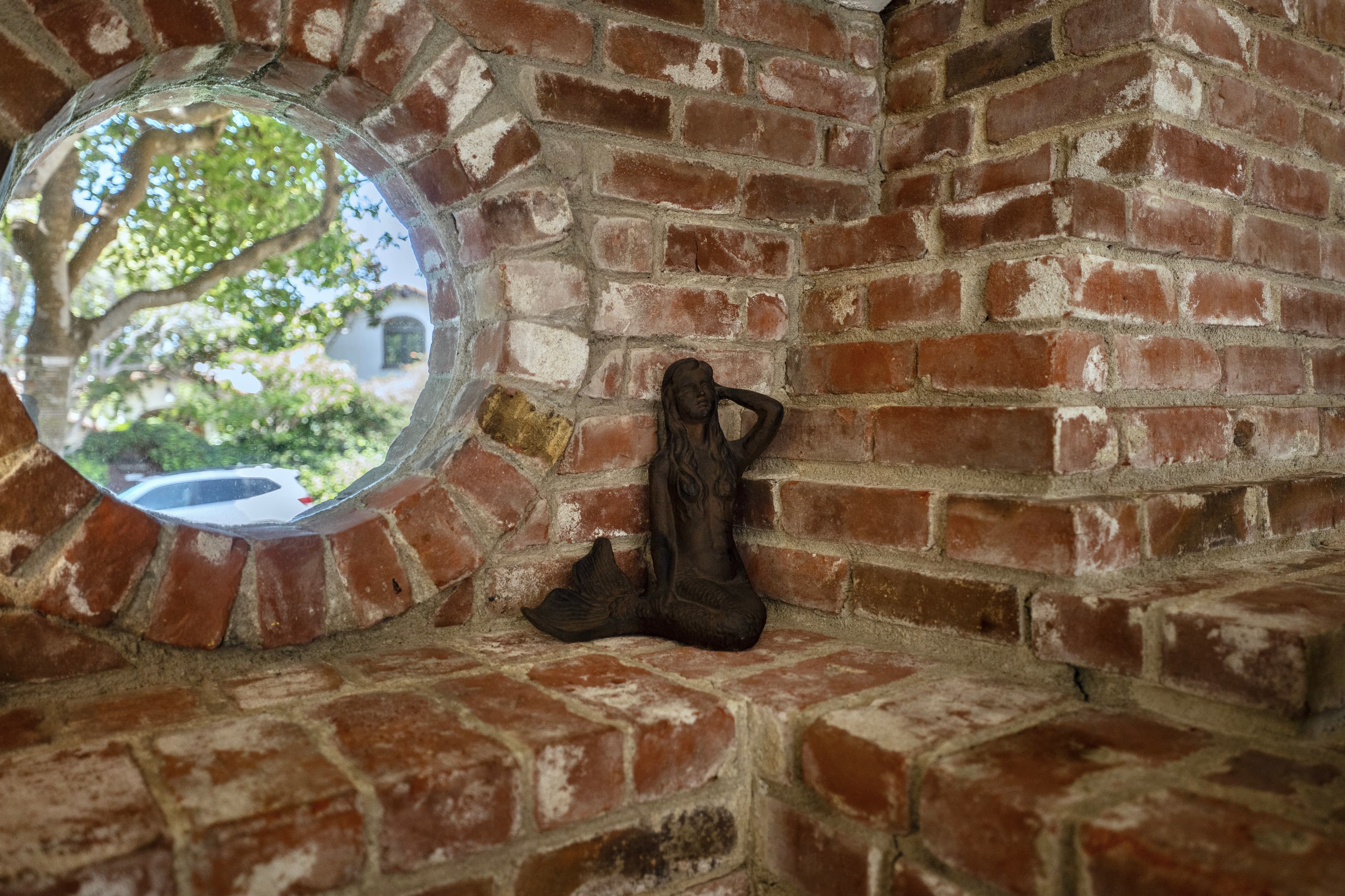 A detailed view of a rustic brick wall featuring a unique circular porthole window and a small bronze mermaid statue resting on the brick ledge.