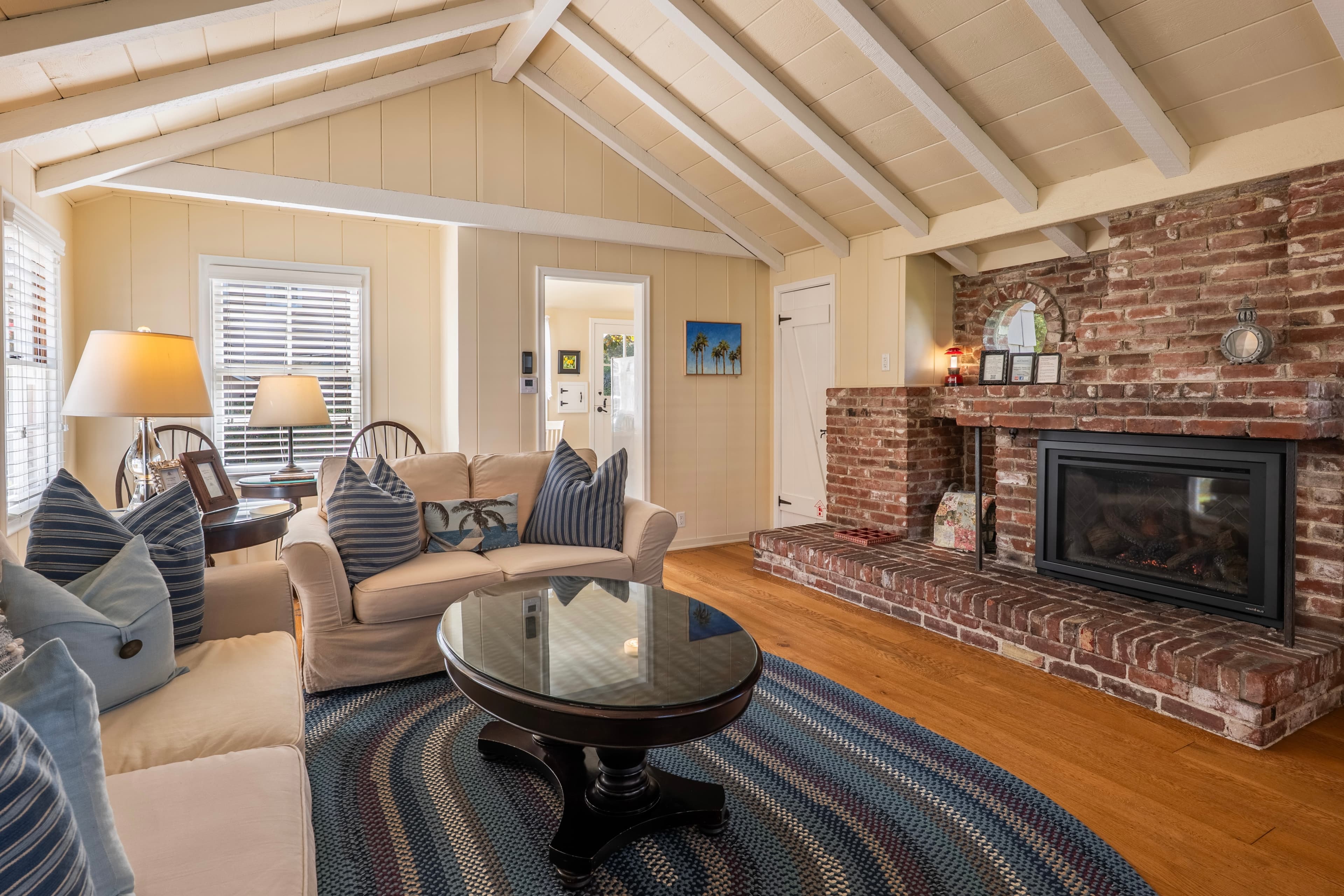 An interior view of a cozy cottage living room featuring a vaulted ceiling with exposed white beams, a large brick fireplace with a black insert, neutral beige sofas with blue accent pillows, and a round dark wood coffee table on a patterned blue rug.