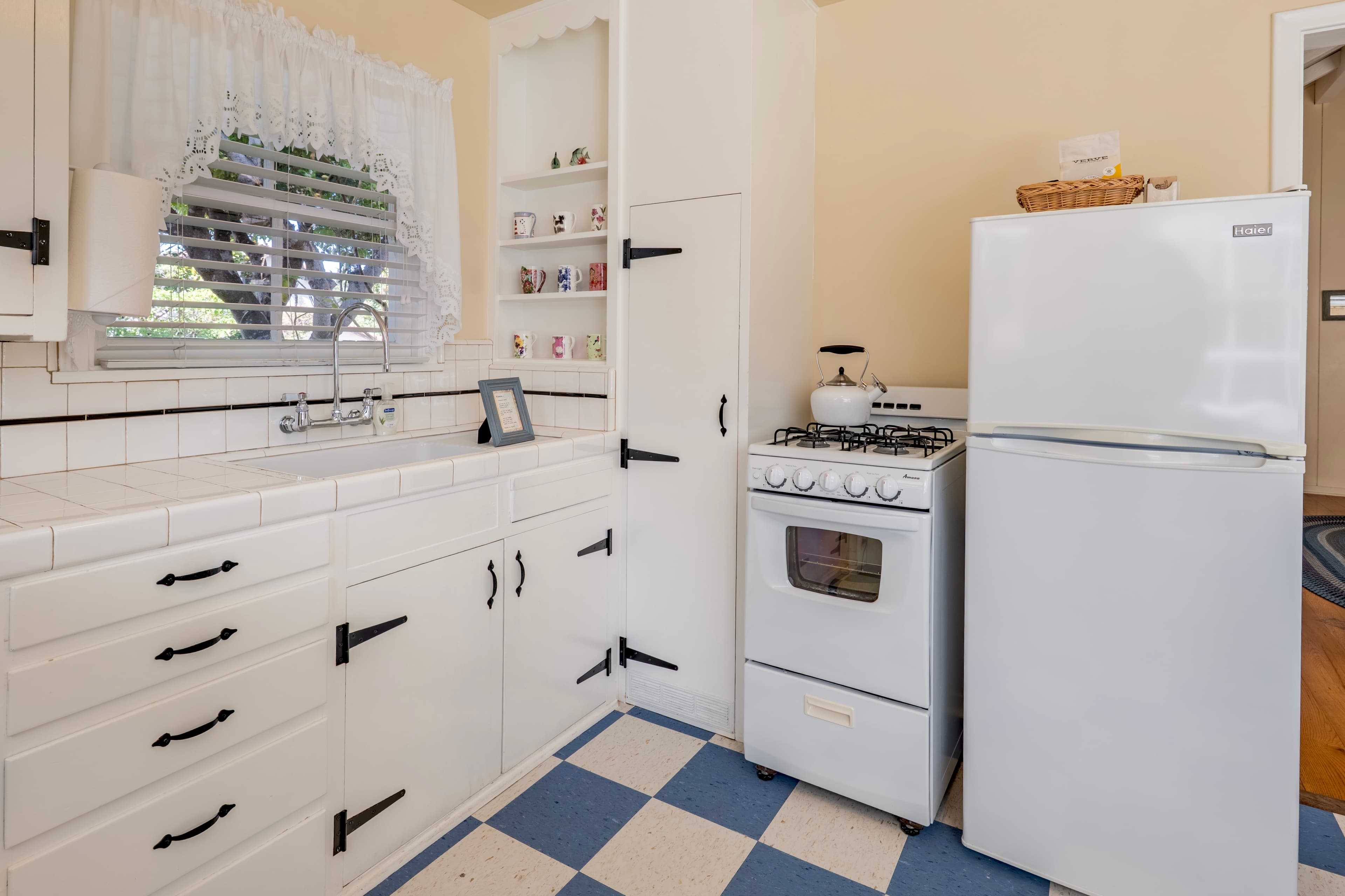 A retro-modern kitchen featuring white cabinetry with vintage black hardware, a white gas stove, and a refrigerator set against a classic blue and white checkered tile floor.