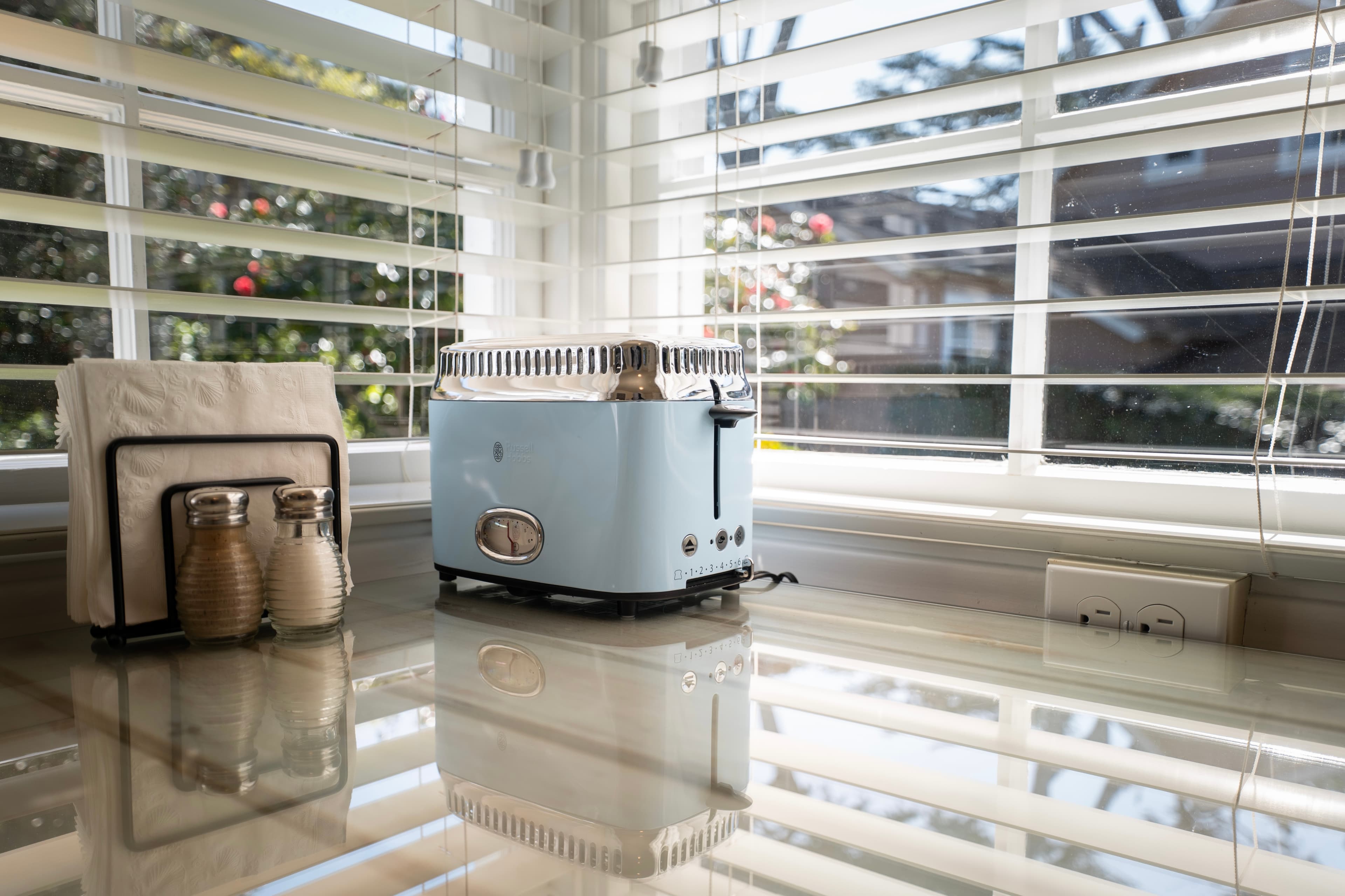 A light blue retro-style toaster sits on a reflective white glass tabletop next to a black napkin holder, positioned in front of a large window with white horizontal blinds.