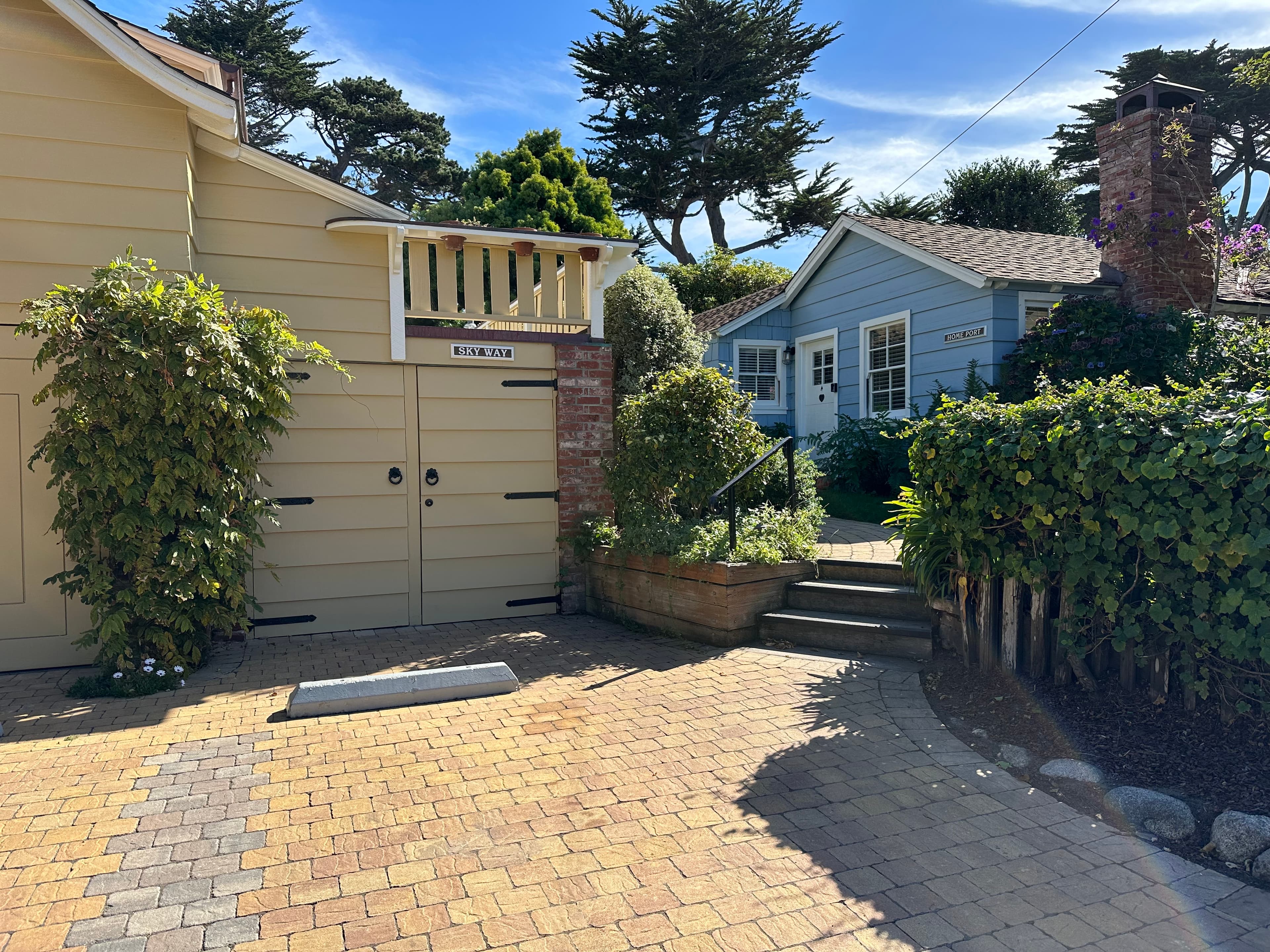 Exterior view of a paved driveway leading to a tan garage and a blue cottage in the background, surrounded by green hedges and tall trees under a clear blue sky.