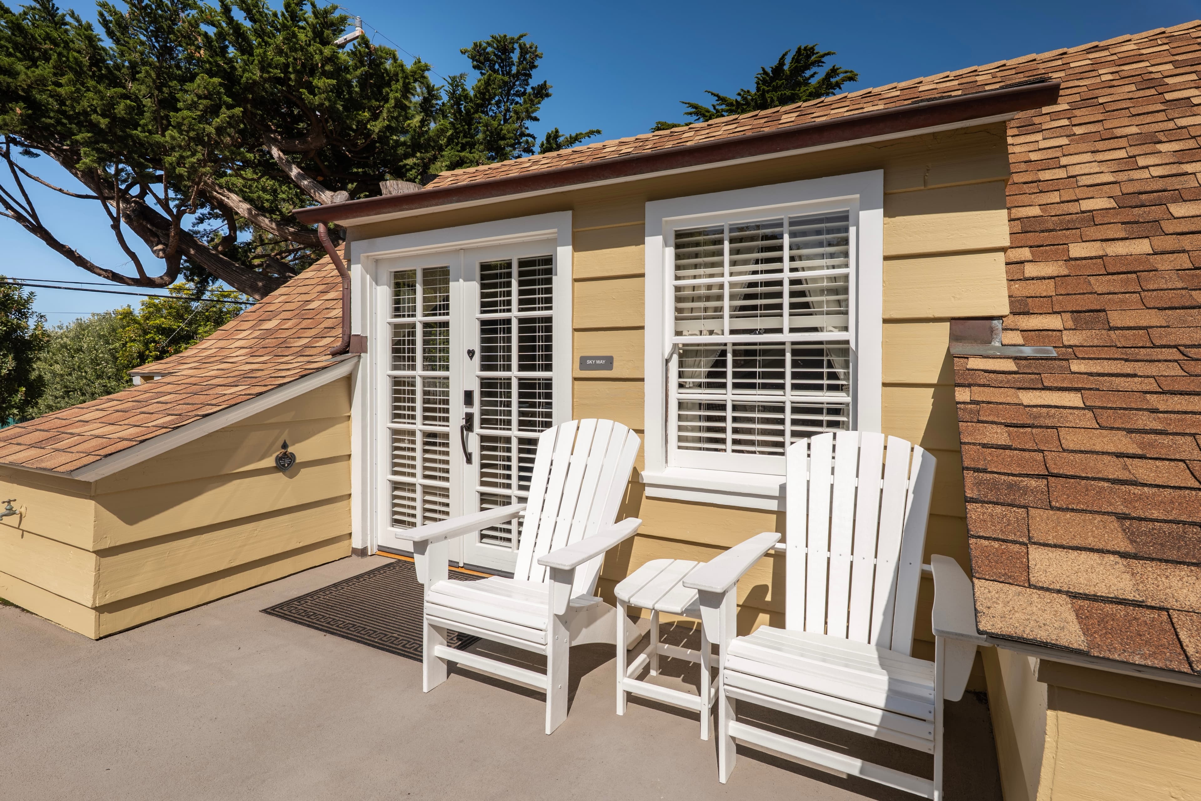 A sunny rooftop balcony with tan siding and a shingled roof, featuring two white Adirondack chairs and a small side table next to a set of white French doors.