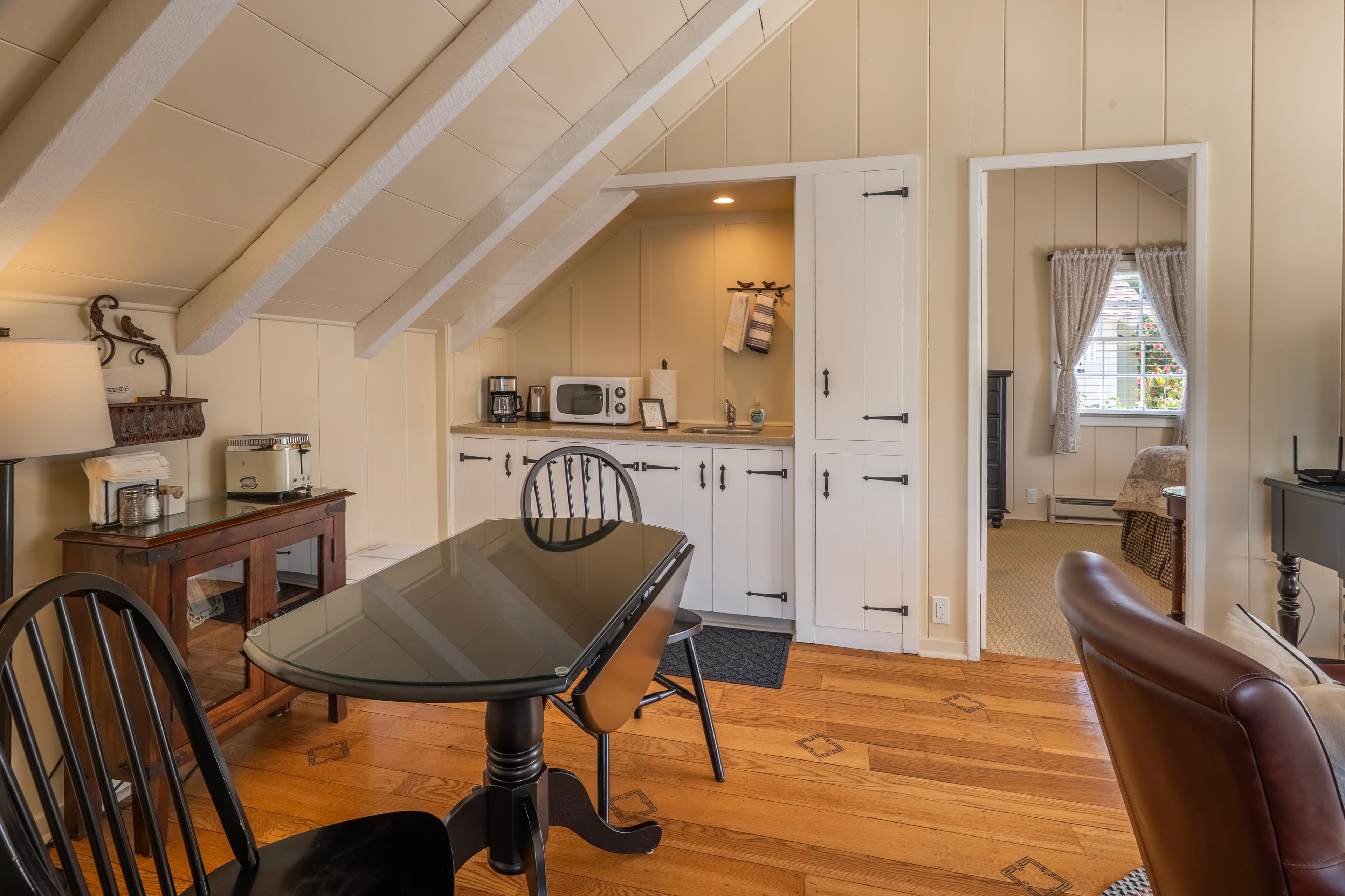 A cozy dining and kitchenette area with vaulted white-beamed ceilings and hardwood floors, featuring a dark wood gate-leg table and a small kitchenette with white cabinets and a microwave.