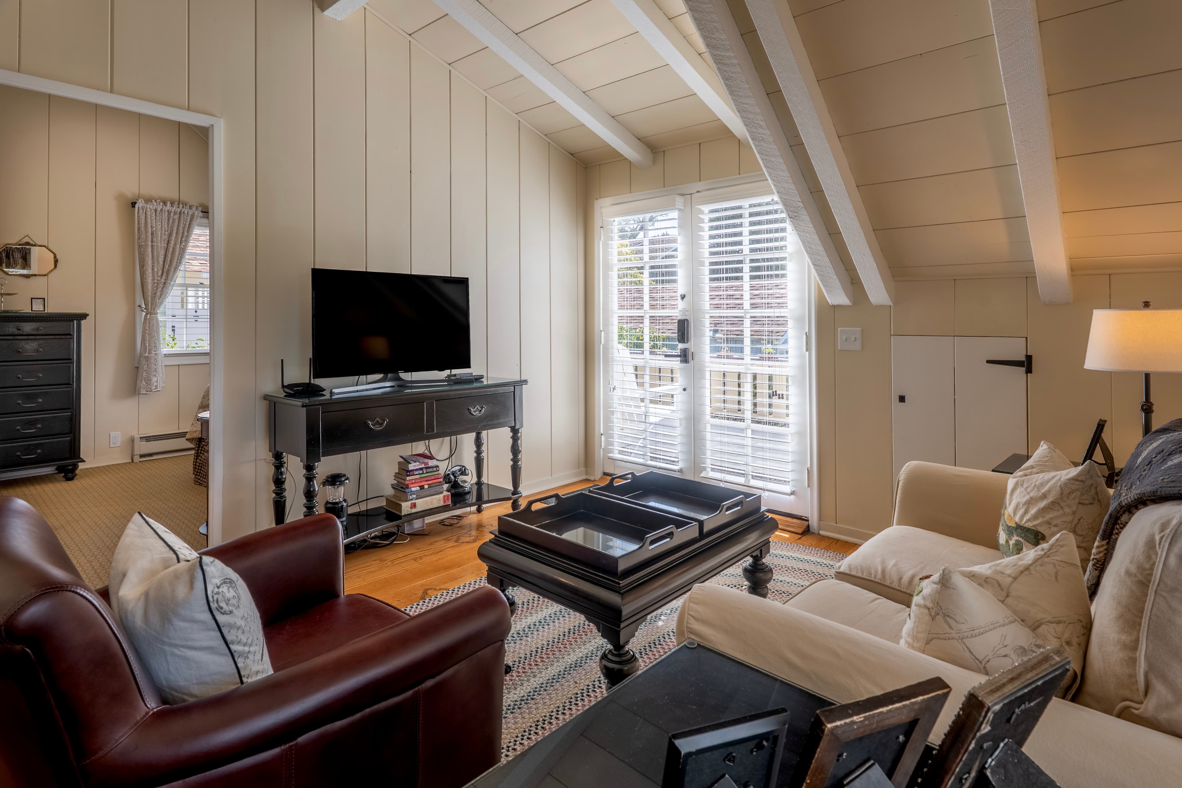 A bright living area with vaulted white-beamed ceilings and hardwood floors, featuring a dark wood media console with a television, neutral-toned seating, and white French doors leading to an outdoor space.