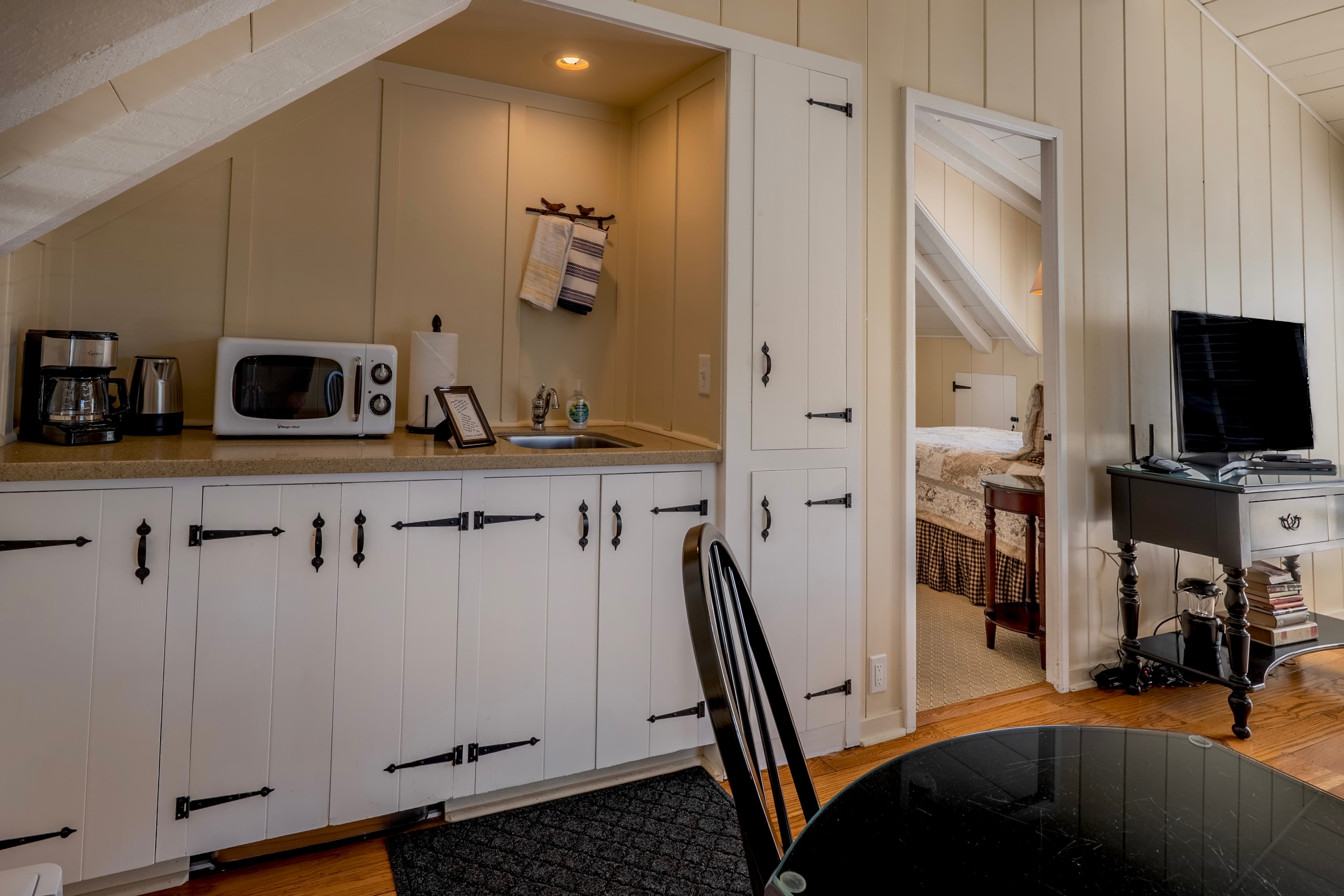 A compact kitchenette in a roof sloped alcove with sloped ceilings, featuring white cabinets with black rustic latches, a small microwave, and a coffee maker on the counter.