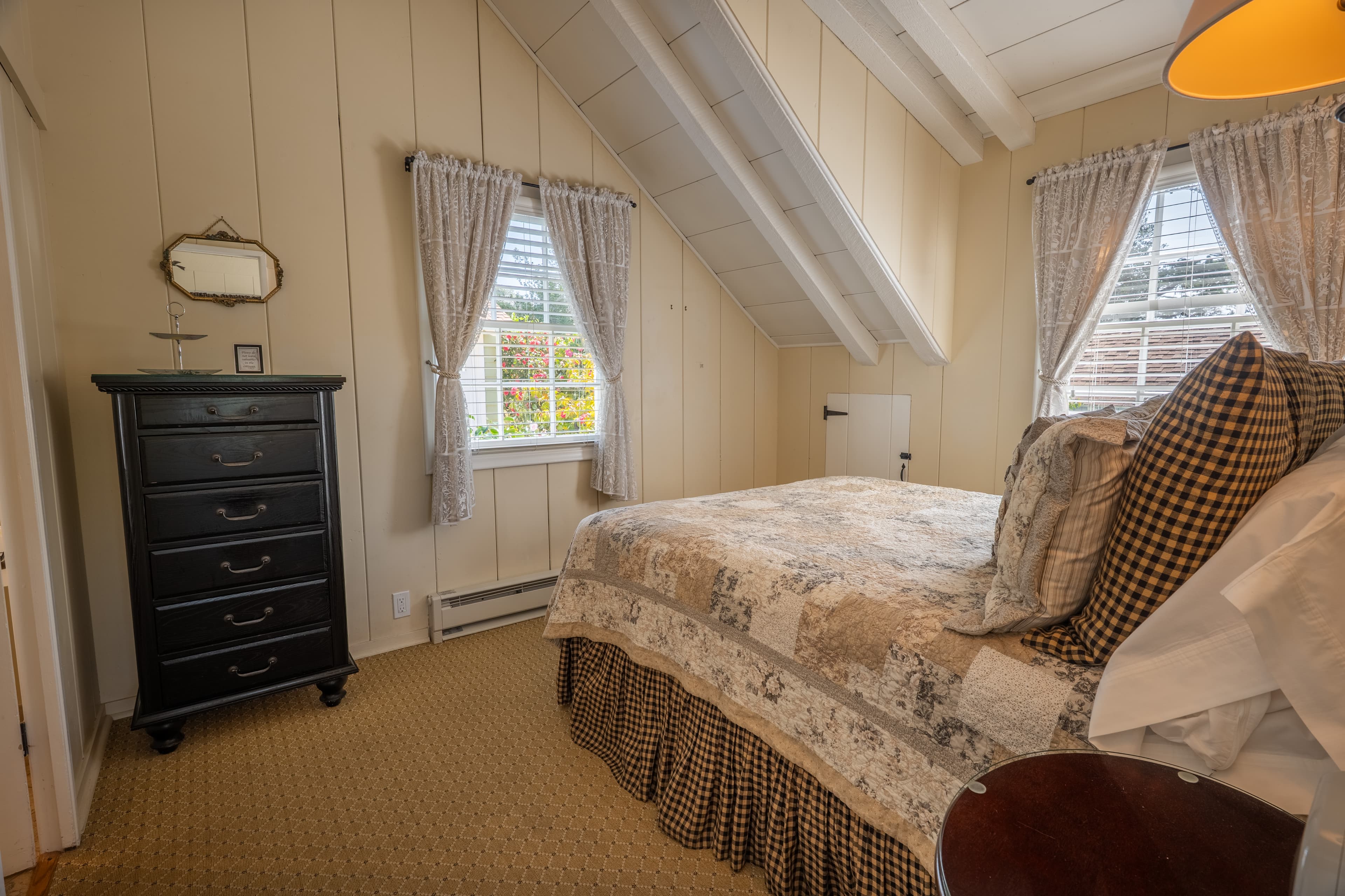A cozy guest bedroom with sloped cream walls and vaulted white-beamed ceilings, featuring a bed with a patterned quilt and a black five-drawer dresser.