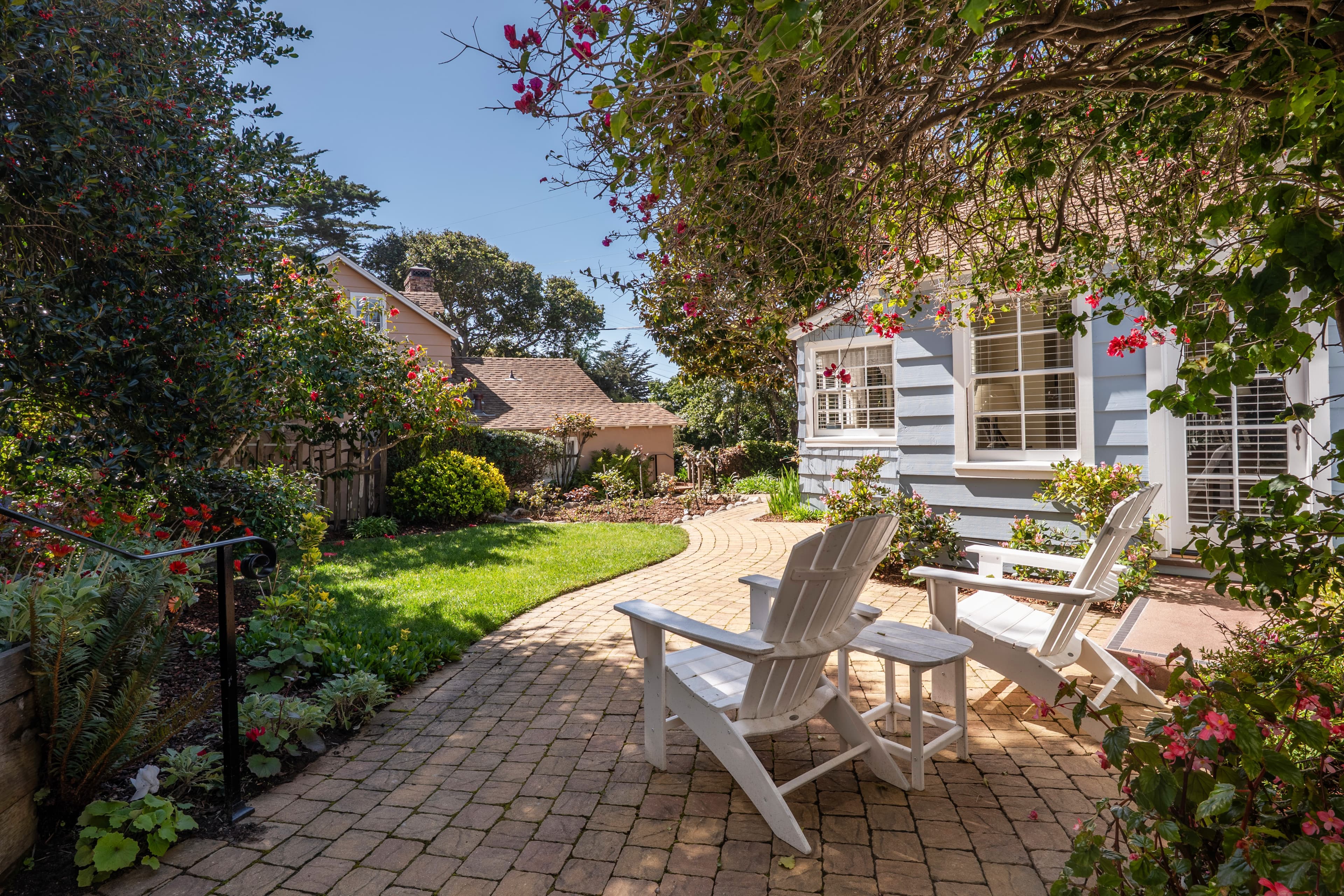 A sunny garden pathway leads to white chairs framed by lush greenery and blooming flowers.