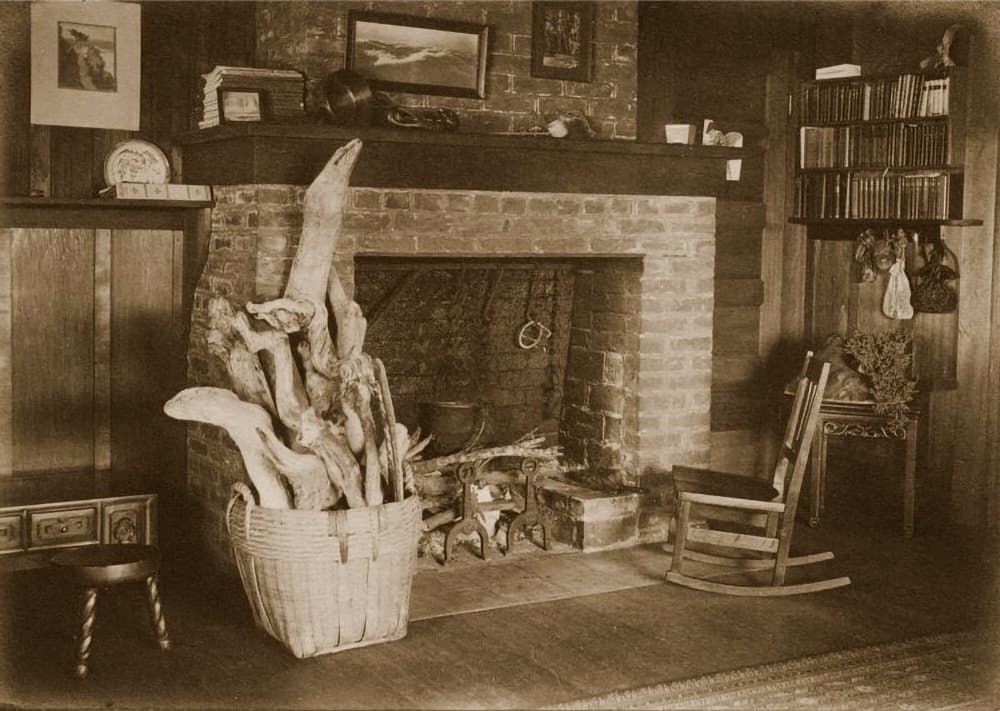 A cozy vintage interior featuring a brick fireplace, a basket of firewood, a rocking chair, and shelves of books.
